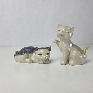 May include: Two ceramic cat figurines with a glossy finish. One cat is lying down with a dark blue and grey patterned back, while the other cat is sitting upright with one paw raised, featuring a solid light grey color and a smooth, reflective surface.