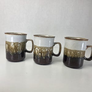Może przedstawiać: Trzy ceramiczne kubki z trójkolorowym szkliwem. Górna część jest nakrapiana w kolorze złamanej bieli, przechodząca w teksturowaną, brązową warstwę środkową z zaciekami, kończącą się solidną, ciemnobrązową podstawą. Każdy kubek posiada wygięte ucho.