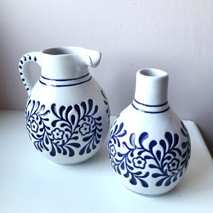 Op de afbeelding: Een witte keramieken kan en een bijpassende vaas, beide met een rond lichaam en een donkerblauw geschilderd bloemmotief. De kan heeft een gebogen handvat en een schenktuit, terwijl de vaas een smalle hals en een ronde opening aan de bovenkant heeft.