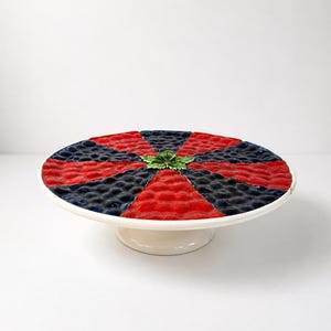 May include: A white ceramic cake stand with a circular top featuring a red and navy blue geometric pattern. A small green leaf is in the center. The stand is elevated on a short, rounded base.