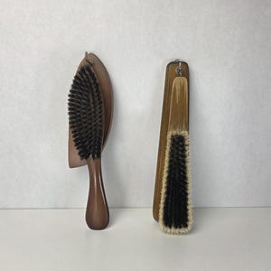 Op de afbeelding: Twee houten borstels met donkere haren. De linkerborstel heeft een ovale kop en een gladde steel. De rechterborstel is rechthoekig met een lichte houten steel en een metalen lus aan de bovenkant om op te hangen. Beide hebben dichte, donkere haren.