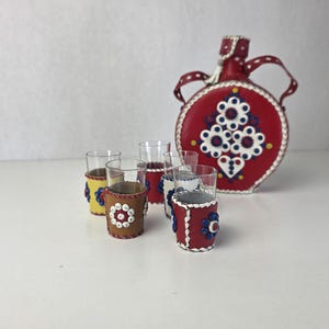 May include: A red leather-covered canteen with a white, blue, and yellow floral pattern and a matching set of five small glass cups, each wrapped in a leather sleeve featuring colourful circular designs in red, blue, yellow, and white.
