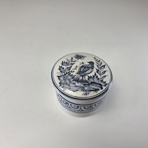 May include: A round ceramic lidded jar featuring a white base with blue hand-painted details. The lid displays a bird perched among floral foliage, while the side of the jar is decorated with a repeating blue geometric pattern around the circumference.