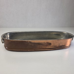 Op de afbeelding: Een ovale koperen bakvorm met een zilverkleurige binnenkant. De buitenkant heeft een warme, gepolijste koperen afwerking met zichtbare gebruikssporen en textuur. Aan beide zijden zijn kleine metalen ringen als handvatten bevestigd.
