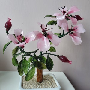 May include: A decorative bonsai tree featuring pink, translucent flowers and green leaves. The flowers have dark pink centers. The tree is planted in a white, square pot filled with small, light-coloured stones.