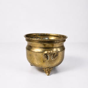 May include: A small, antique brass pot with three ornate feet and two handles. The pot has a rounded body and a wide rim. The brass has a warm, golden color with some areas of darker patina, suggesting age and use. The pot is likely decorative.