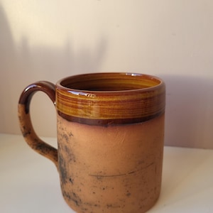 May include: A cylindrical ceramic mug with a matte, light brown, speckled base and a glossy, dark brown glazed rim. The mug features a matching curved handle with dark brown glaze accents and a smooth, polished interior finish.