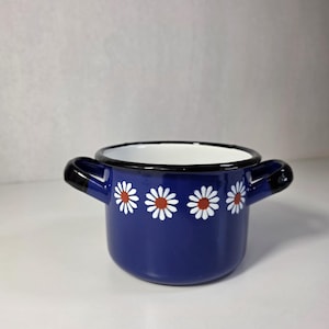 Op de afbeelding: Een kleine, ronde, diepblauwe emaille pan met een wit en oranje madeliefjespatroon aan de buitenkant. De pan heeft twee zwarte handvatten aan de zijkanten, een zwarte rand, een glanzende afwerking en een witte binnenkant.