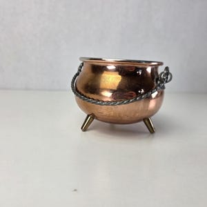 Puede incluir: Un pequeño caldero de metal color cobre con acabado pulido. El recipiente cuenta con tres patas cortas y cónicas, y un asa de metal trenzado en tono plateado sujeta a los lados con pequeñas anillas. El borde es liso y metálico.