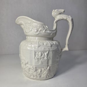 Może przedstawiać: Kremowy dzbanek ceramiczny z ozdobnym uchwytem zwieńczonym rzeźbioną figurką zwierzęcia. Korpus dzbanka jest ozdobiony szczegółowymi wzorami reliefowymi, w tym motywami kwiatowymi i elementami architektonicznymi wokół górnej krawędzi.