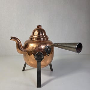 Op de afbeelding: Een koperen theepot met een ronde vorm, een bijpassend deksel en een gebogen tuit. De theepot heeft drie donkere metalen poten bevestigd met driehoekige platen en een lange, conische, zilverkleurige metalen handgreep aan de zijkant.