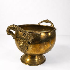 Könnte beinhalten: Eine Vintage-Messingschale mit zwei verzierten Griffen und einem Sockel. Die Schale hat einen goldenen Farbton und weist detaillierte Reliefarbeiten auf, darunter ein Gesicht an der Seite. Die Griffe haben ein strukturiertes Design. Diese dekorative Schale ist ideal für die Inneneinrichtung.