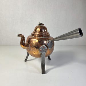Könnte beinhalten: Eine kupferne Teekanne mit rundem Körper, einem kleinen gebogenen Ausguss und einem langen, spitz zulaufenden silberfarbenen Griff. Die Teekanne hat einen gewölbten Deckel mit einem kleinen Knauf und steht auf drei stabilen, flachen silberfarbenen Beinen.