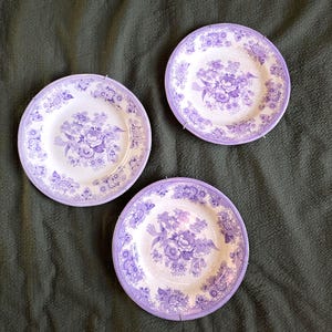 Puede incluir: Tres platos de cerámica blanca con un diseño floral púrpura. Cada plato presenta un ramo central de flores y un borde de patrones florales más pequeños. Los platos están dispuestos sobre una superficie verde oscuro.