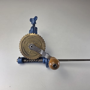 Può includere: Un trapano a mano in metallo blu con un grande ingranaggio in ottone con segni visibili, una manovella in metallo argentato e un'impugnatura in legno liscia e arrotondata. L'utensile include un meccanismo a barra filettata e una vite a farfalla.