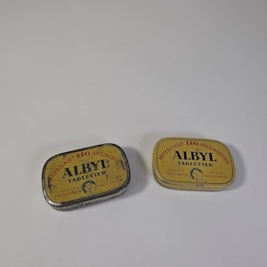 May include: Two rectangular metal tins with rounded corners, featuring yellow and black labels. The text reads Aktiebolaget Leo Hälsingborg, Albyl Tabletter, and includes a small circular logo with a lion head. One tin shows signs of wear and age.