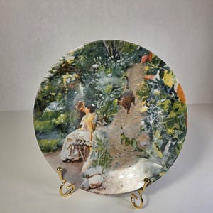 Peut inclure: Une assiette décorative ronde en céramique ornée d'une peinture impressionniste colorée représentant une personne assise sur un banc dans un jardin luxuriant avec un chat marchant à proximité. L'assiette repose sur un support en métal doré.