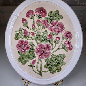 Può includere: Un piatto decorativo in ceramica bianca con un motivo floreale in rilievo. I fiori sono di un rosa intenso con foglie e steli verdi. Il piatto ha un centro color crema e un bordo bianco. Il piatto è esposto su un supporto dorato.