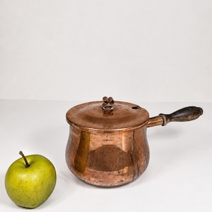 Pode incluir: Uma panela de cobre com tampa e cabo de madeira. A panela tem um corpo arredondado e um pequeno botão na tampa. Uma maçã verde está à esquerda da panela. A panela é provavelmente vintage.