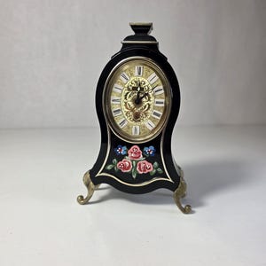 May include: A black mantel clock with a glossy finish, featuring an oval face with Roman numerals and gold-toned filigree detailing. The lower section is decorated with a hand-painted floral motif of pink roses and blue flowers, supported by three ornate gold-toned metal feet.