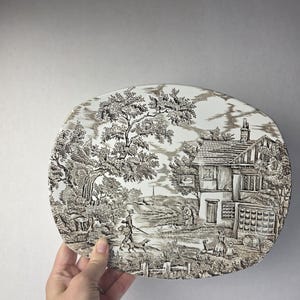 Op de afbeelding: Een ovale keramische schaal met een bruin-witte transferware-scène van een landelijk Engels dorp. De illustratie toont een herberg met het opschrift The Swan, een persoon die met een hond wandelt, bomen en figuren in een landschap.