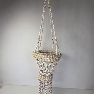 Può includere: Un supporto per piante sospeso costruito con una rete di conchiglie bianche e marroni. La struttura presenta un bordo circolare decorato con conchiglie, un corpo a rete intrecciato fatto di piccole conchiglie e lunghi fili pendenti di conchiglie.