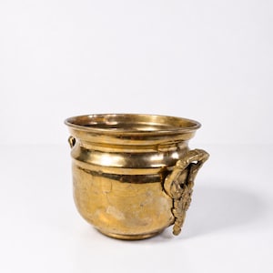 Puede incluir: Una jardinera de latón vintage con un cuerpo redondeado y asas ornamentadas. La jardinera tiene un borde ancho y una superficie ligeramente texturizada. Las asas presentan un diseño detallado, posiblemente floral. El color general es un oro cálido y envejecido.