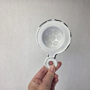 Może przedstawiać: Białe sitko do herbaty z emaliowanego metalu z okrągłą miseczką z perforowanym wzorem małych otworów. Krawędzie obręczy i uchwytu wykazują postarzane czarne detale, a uchwyt posiada okrągły otwór do zawieszenia lub przechowywania.
