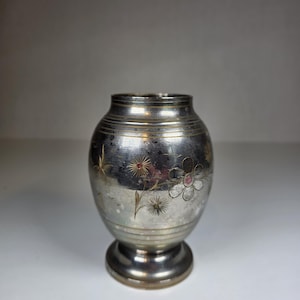May include: A small, silver-toned vase with a rounded body and a pedestal base. The vase is adorned with etched floral designs, including a large flower with a pink center and smaller starburst-like flowers. The metal has a slightly tarnished appearance.