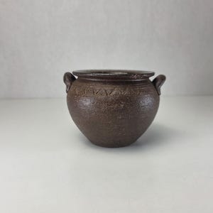 Peut inclure: Un petit pot en céramique brun foncé, de forme arrondie, avec une surface texturée et mouchetée. Le pot présente deux petites anses sur les côtés opposés et une bande décorative de motifs triangulaires incisés autour du corps.