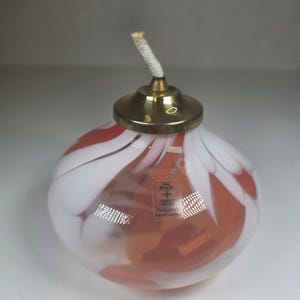 Puede incluir: Una lámpara de aceite redonda de vidrio con tapa y mecha de latón. La lámpara presenta un diseño de remolino blanco y rojo. La etiqueta dice "Exclusive hand-crafted".