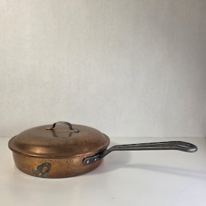 May include: A copper-colored frying pan with a lid and a long, dark handle. The pan has a rounded shape and a slightly raised rim. The handle is riveted to the pan. The lid has a small handle. The pan is likely vintage.