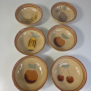 May include: A set of six round ceramic bowls with a cream-coloured base and brown striped rims. Each bowl features a hand-painted fruit illustration at the centre, including bananas, grapes, a melon, an apple, a peach, and a pair of cherries.