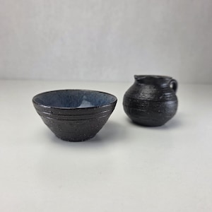 Puede incluir: Un cuenco pequeño de cerámica oscura y texturizada con un interior liso de color azul claro junto a una jarra de leche de cerámica oscura y texturizada a juego. Ambas piezas presentan un acabado exterior mate y rugoso con ranuras horizontales.