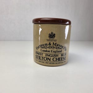May include: A cylindrical beige ceramic jar with a dark brown lid. The front features black text reading Fortnum & Mason Plc London England Finest English Blue Stilton Cheese, along with a crest and the text 16 oz. KEEP COOL BEST BEFORE.