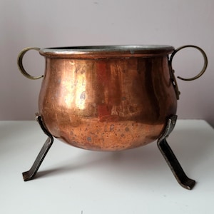 Könnte beinhalten: Ein kleiner, antiker Kupferkessel mit Messinggriffen und einem silberfarbenen Rand. Der Kessel steht auf einem dreibeinigen, dunklen Metallständer. Das Kupfer hat einen warmen, rotbraunen Farbton mit sichtbarer Patina.