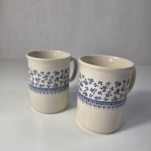 Peut inclure: Deux tasses en céramique blanc cassé ornées d'un motif floral bleu sur la partie supérieure. Chaque tasse présente un corps cylindrique, une anse lisse sur le côté et une bordure décorative bleue en forme de dentelle sous les fleurs.