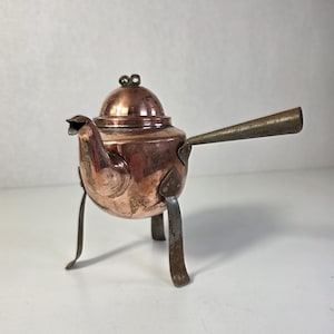 Op de afbeelding: Een kleine, antieke koperen theepot met een deksel en een handvat. De theepot heeft een tuit en staat op drie metalen poten. Het koper heeft een warme, roodbruine kleur en het handvat is van donkerder, contrasterend metaal. De theepot is waarschijnlijk vintage.