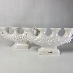 Può includere: Due candelabri in ceramica bianca con un design a più candele e motivi floreali in rilievo sulla parte anteriore. Ogni pezzo ha una base larga e piccole maniglie ad anello sui lati. È visibile una piccola etichetta con la scritta Rose Spray.