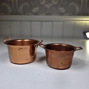 Può includere: Due pentole di rame con piccole maniglie. Le pentole hanno una texture martellata e una finitura lucida. Le pentole sono di diverse dimensioni, la più grande a sinistra e la più piccola a destra. Sono su una superficie bianca.