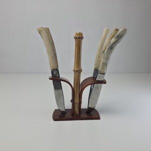 May include: A set of three knives with curved, light-colored textured handles held in a dark brown wooden stand. The stand features a central vertical bamboo-style post and two curved side supports that hold the knives in an upright position.