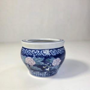 May include: A small, round ceramic planter with a dark blue background and white trim. The planter features a floral design with pink flowers and green leaves. The rim has a white and blue heart pattern.