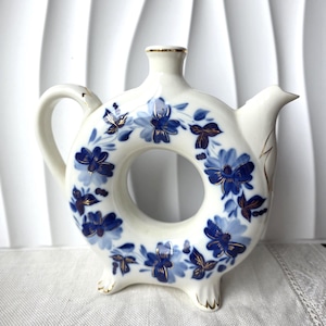 Könnte beinhalten: Eine weiße Keramik-Karaffe mit rundem Körper, Henkel, Ausguss und drei kleinen Füßen. Sie ist mit blauen Blumenmustern und goldenen Akzenten verziert. Das Design erinnert an Vintage-Porzellan.