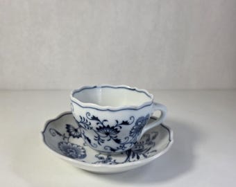Pode incluir: Um conjunto de chávena de chá e pires em porcelana branca com rebordo recortado e um delicado padrão floral azul. O design inclui motivos botânicos intrincados e vinhas pintadas num tom azul profundo sobre a superfície de cerâmica.