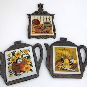 Puede incluir: Tres salvamanteles decorativos vintage. Uno es rectangular con una escena de cocina, y dos tienen forma de tetera. Un salvamanteles de tetera tiene flores amarillas, y el otro tiene un diseño de barco y casco vikingo. Todos tienen un marco de metal oscuro.