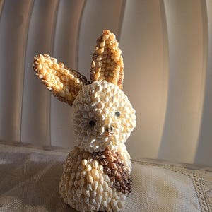 May include: A decorative bunny sculpture crafted from small seashells. The bunny features long ears, a rounded body, and a small, dark brown patch on its chest. The sculpture is mainly cream-coloured with tan and brown accents.