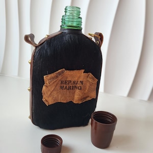 Può includere: Una borraccia rivestita in pelliccia nera con una toppa in pelle marrone recante la scritta REP. SAN MARINO. La borraccia presenta un collo in vetro verde, borchie metalliche dorate sul lato e due tappi in plastica marrone separati davanti.