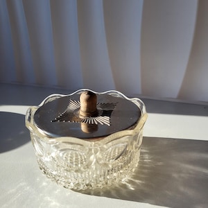 Op de afbeelding: Een heldere glazen pot met een zilveren deksel en een houten knop. De pot heeft een decoratief patroon rond de zijkanten en een stervormig ontwerp op het deksel. De pot staat op een wit oppervlak.