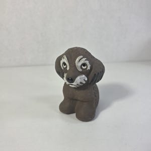 May include: A small, brown ceramic dog figurine with white detailing around the eyes and muzzle. The dog is sitting upright with its ears perked up. The figurine has a smooth, matte finish.