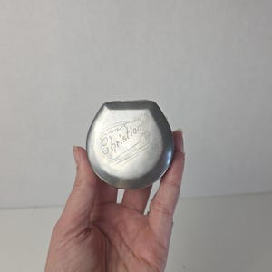 May include: A small, round, silver-toned metal pill box with a hinged lid. The top of the lid features an engraved design of a feather and the name "Christian" in cursive script. The metal surface has a smooth, matte finish with visible metallic sheen.